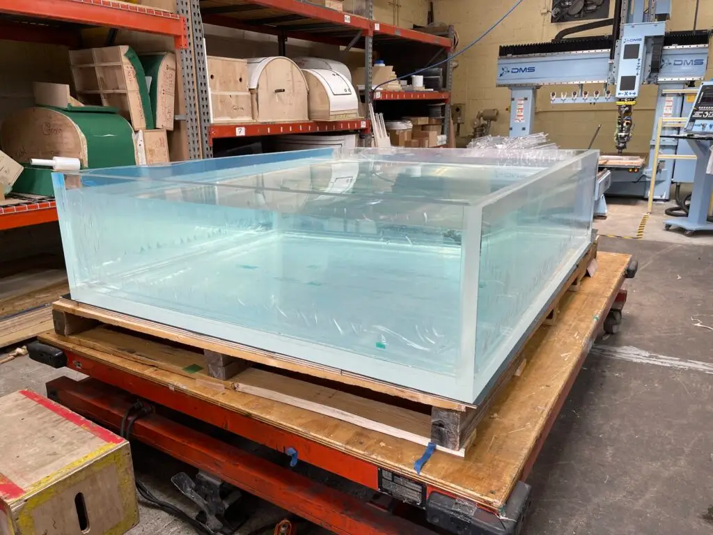 A large clear acrylic tank built by Plastic-Craft on a shop cart, with a DMS CNC router and material racks in the background of the West Nyack fabrication shop